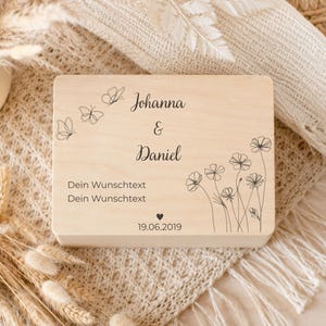 Eine hellholzfarbene Andenkenbox mit der Gravur „Johanna & Daniel Dein Wunschtext Dein Wunschtext 19.06.2019“ sowie Schmetterlings- und Blumenmotiven, auf einem cremefarbenen Untergrund mit getrockneten Gräsern.