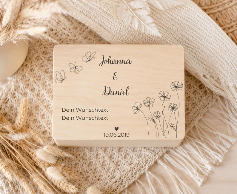 Eine hellholzfarbene Andenkenbox mit der Gravur „Johanna & Daniel Dein Wunschtext Dein Wunschtext 19.06.2019“ sowie Schmetterlings- und Blumenmotiven, auf einem cremefarbenen Untergrund mit getrockneten Gräsern.