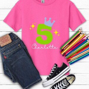 May include: A pink t-shirt with a green number 5 and a blue crown design. The name "Charlotte" is written in cursive below the number. The shirt is paired with blue jeans and black sneakers. Colored pencils are also visible.