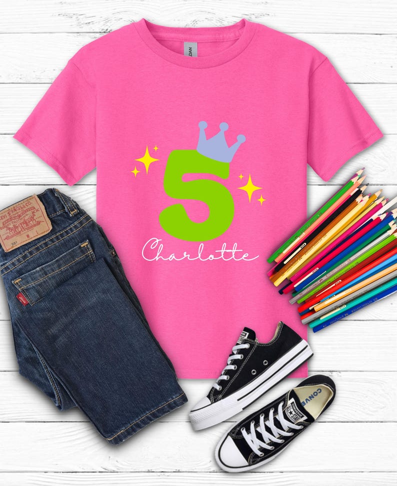 May include: A pink t-shirt with a green number 5 and a blue crown design. The name "Charlotte" is written in cursive below the number. The shirt is paired with blue jeans and black sneakers. Colored pencils are also visible.