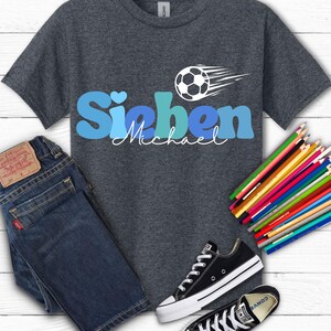 May include: Dark gray t-shirt with the name "Sieben" in blue letters and the name "Michael" in white script. A soccer ball graphic with motion lines is above the name. The image also includes blue jeans, black sneakers, and colored pencils.