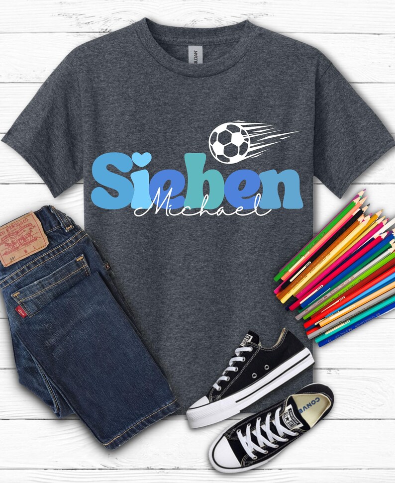 May include: Dark gray t-shirt with the name "Sieben" in blue letters and the name "Michael" in white script. A soccer ball graphic with motion lines is above the name. The image also includes blue jeans, black sneakers, and colored pencils.