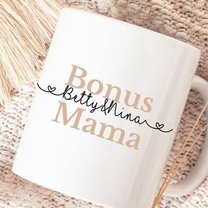 Puede incluir: Taza de cerámica blanca con las palabras "Bonus Mama" en color beige, con "Betty & Nina" en escritura cursiva. Una línea negra con dos corazones separa las palabras. La taza tiene un asa curva.