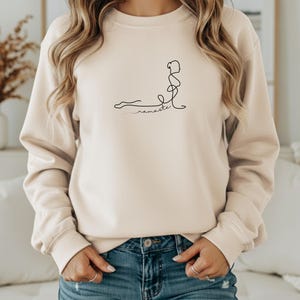 May include: A cream-coloured sweatshirt featuring a black line drawing of a person in a yoga pose. The word "namaste" is written in cursive below the figure. The sweatshirt has long sleeves and a crew neck.