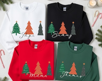 Puede incluir: Cuatro sudaderas de cuello redondo en blanco, negro, rojo y verde. Cada una presenta un diseño de árbol de Navidad con un árbol verde, naranja y gris, y una estrella dorada. La sudadera blanca dice "Papa", la negra dice "Sammy", la roja dice "Mama" y la verde dice "Thomas".