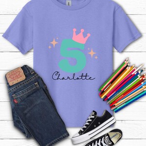 May include: A lilac-colored t-shirt with a teal number 5, a pink crown, and the name "Charlotte" in black script. The image also includes blue jeans, black sneakers, and a set of colored pencils, all arranged on a white wooden surface.