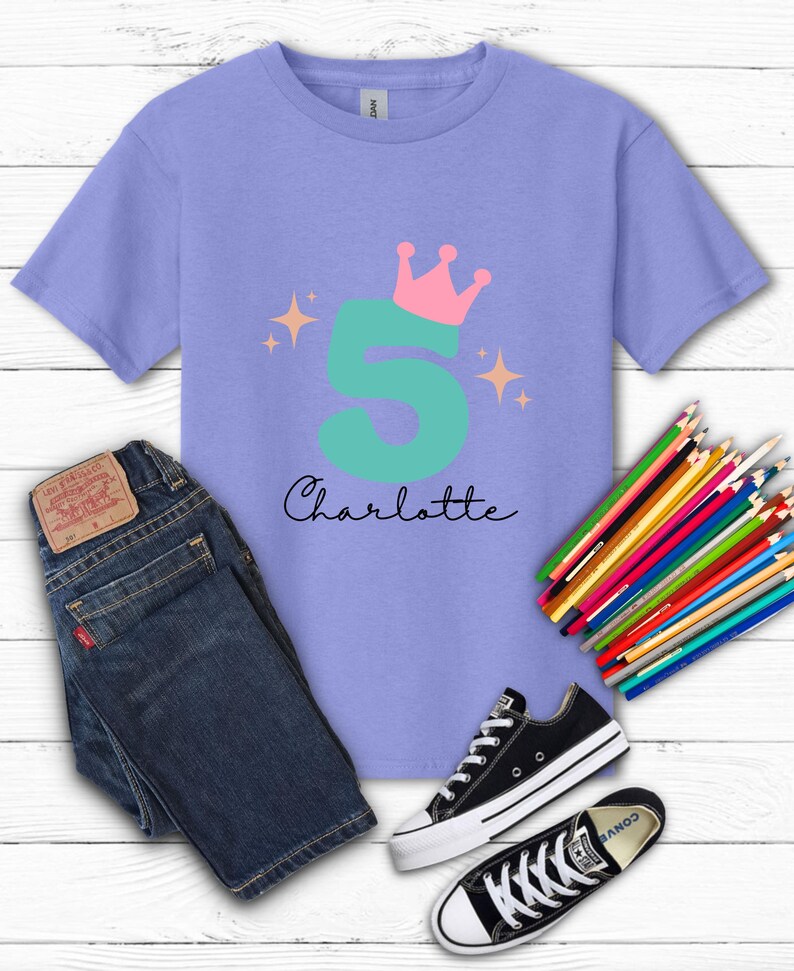 May include: A lilac-colored t-shirt with a teal number 5, a pink crown, and the name "Charlotte" in black script. The image also includes blue jeans, black sneakers, and a set of colored pencils, all arranged on a white wooden surface.