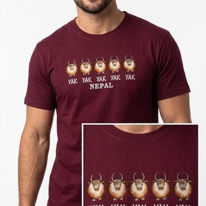 Pode incluir: T-shirt bord&ocirc; com um desenho bordado de cinco iaques e o texto "YAK YAK YAK YAK YAK NEPAL" em branco. A camisa &eacute; de manga curta e tem gola redonda.