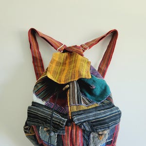 May include: A colorful patchwork backpack with adjustable straps. The backpack features a variety of fabrics in shades of red, yellow, blue, and green. It has a drawstring closure and two front pockets with wooden toggles.
