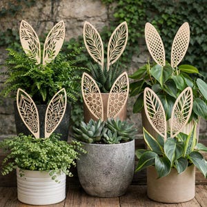 Op de afbeelding: Houten plantenstekers in de vorm van konijnenoren met ingewikkelde uitgesneden patronen. De stekers worden in diverse potplanten geplaatst, waaronder varens en vetplanten. De konijnenoren hebben verschillende patronen.