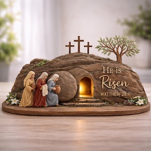 May include: A religious Easter scene depicts three women approaching an open tomb. The tomb is carved into a rock formation, with three crosses and a tree on top. The words "He is Risen" and "Matthew 28:6" are inscribed on the rock.