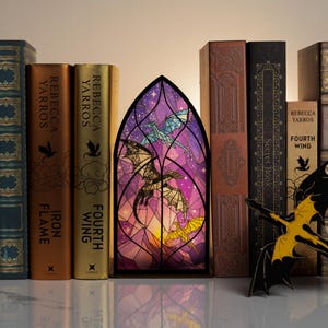 May include: A decorative stained glass window with a dragon design, set between a collection of books. The books include titles like "Fourth Wing" and "Iron Flame" by Rebecca Yarros. A black and yellow dragon figurine is in front.