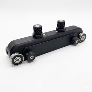 May include: A black, elongated mechanical component featuring a textured finish, equipped with four silver-toned ball bearings at the corners and two prominent, knurled black adjustment knobs positioned along the top surface.