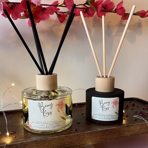 May include: Two reed diffusers, one clear glass and one black, with wooden tops. The clear diffuser has black reeds, the black one has natural reeds. Both feature "Berry Kiss" labels with floral designs. Pink flowers and foliage are in the background.