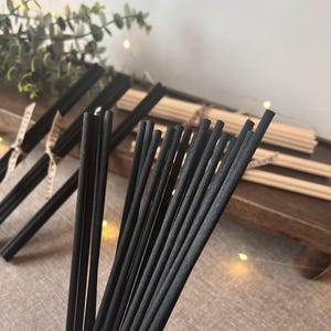 May include: Close-up of black diffuser reeds, bundled and arranged. Several bundles of black reeds are in focus, with additional bundles of natural-coloured reeds in the background. The reeds are on a wooden surface, with greenery and lights.