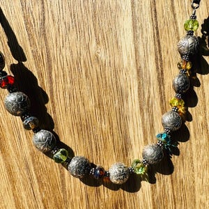 May include: A necklace featuring a series of round, etched silver-toned metal beads interspersed with smaller, translucent faceted glass beads in vibrant shades of red, yellow, green, and blue, connected by a dark metal chain.