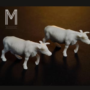 May include: Two white miniature cow figurines. The cows are in a walking pose, with visible horns and detailed body features. The background is a dark brown colour. The text "M" and "MAX MINIATURES" is in the upper left corner.