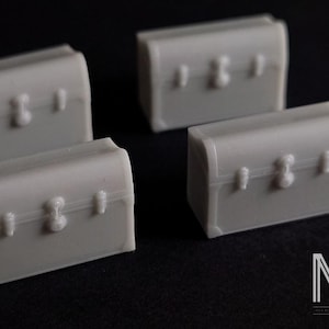 May include: Four miniature, pale grey treasure chests with curved lids and decorative fittings are arranged on a dark background. The chests feature a traditional design with a central clasp and side details. The image has studio lighting.