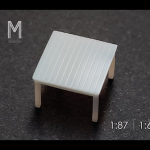 May include: A small, white miniature table with a slatted top and four legs. The table is designed for a scale model, with the text "1:87 | 1:64" visible at the bottom. The table is approximately 1 inch square.