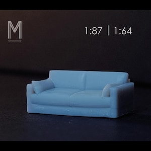 Pode incluir: Um sofá em miniatura azul claro com duas almofadas e apoios de braços. O sofá é um modelo para projetos em escala 1:87 e 1:64. O sofá está sobre um fundo escuro.
