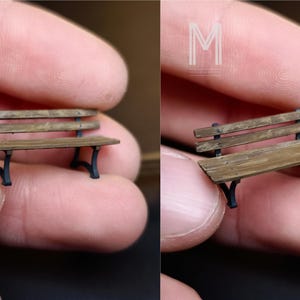 May include: Miniature wooden park bench with a distressed finish. The bench has a slatted seat and backrest, supported by black metal legs. A small-scale model, perfect for a dollhouse or diorama.