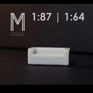 May include: A small, white miniature sofa with a rectangular shape. The sofa has a simple design with armrests and a seat. The image also includes the text "M" and "MAX MINIATURES", along with the scales 1:87 and 1:64.