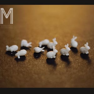 Peut inclure: Plusieurs petites figurines de lapins blancs sur une surface marron. Les lapins ont des détails visibles, comme les oreilles et les queues. Le texte "MAX MINIATURES" avec un logo "M" stylisé est également présent.