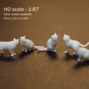 Op de afbeelding: Vijf kleine, witte, onbeschilderde kattenbeeldjes in verschillende houdingen. De tekst op de afbeelding luidt: M, HO scale - 1:87, other scales available from 1:12 to 1:160. De katten zijn afgebeeld in staande, zittende en hurkende posities met gladde oppervlakken.