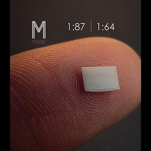 Peut inclure: Un petit objet rectangulaire de couleur claire est posé sur un doigt. L'image comprend le texte "1:87 | 1:64" et un logo "M" stylisé. La texture de l'objet est visible, suggérant une maquette ou un composant miniature.