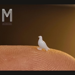 Può includere: Una minuscola statuetta di colomba bianca posata sulla punta di un dito. L'uccello in miniatura è dettagliato con ali e zampe visibili. Lo sfondo è marrone sfocato, con il testo "M" e "MAX MINIATURES" nell'angolo in alto a sinistra.