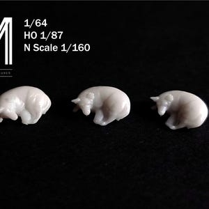 Pode incluir: Três pequenas figuras brancas de ursos polares em miniatura em posição de dormir. As figuras estão dispostas em fila sobre um fundo preto. O texto no canto superior esquerdo diz "1/64 HO 1/87 N Scale 1/160".