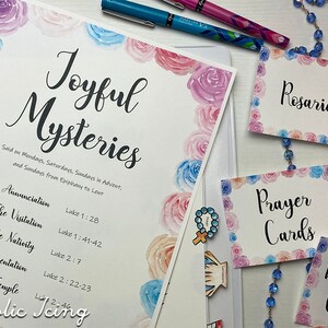 Op de afbeelding: Een verzameling religieuze items, waaronder een document getiteld "Joyful Mysteries" met bloemaccenten, pennen en kaarten met de tekst "Rosaries", "Prayer Cards" en "Baptism Candles". Er is ook een blauwe rozenkrans aanwezig.