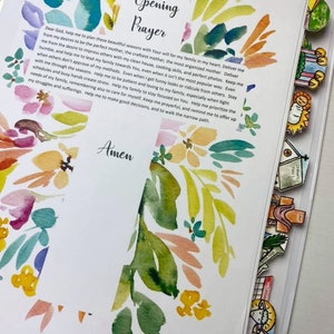 Op de afbeelding: Open map met een aquarel bloemenontwerp rond de tekst "Opening Prayer" en "Amen". De pagina bevat een gebed en is versierd met kleurrijke tabbladen. De woorden "Catholic Icing" staan onderaan.