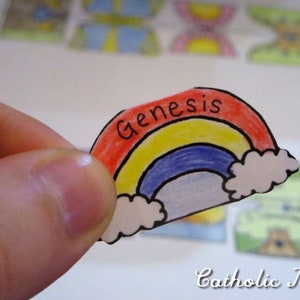 May include: A hand holds a paper cutout of a rainbow with the word "Genesis" written on it. The rainbow is colored red, yellow, blue, and white.