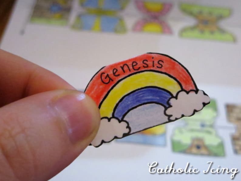 May include: A hand holds a paper cutout of a rainbow with the word "Genesis" written on it. The rainbow is colored red, yellow, blue, and white.