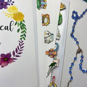 Op de afbeelding: Een planner met ge&iuml;llustreerde tabbladen en bloemaccenten. De plannerpagina's zijn wit, met een bloemenkransontwerp in paars en geel. Een blauwe rozenkrans en pen zijn ook zichtbaar.