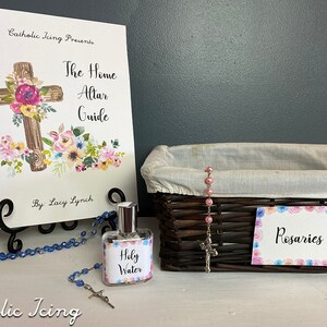 Op de afbeelding: Een Home Altar Guide-boek met een bloemenkruisontwerp, een fles Heilig Water, een rozenkrans en een mand met het label "Rozenkransen". Het boek staat op een zwarte metalen standaard. De items zijn op een wit oppervlak geplaatst.