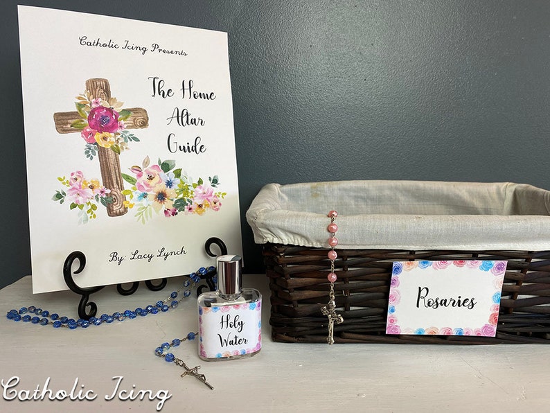 Op de afbeelding: Een Home Altar Guide-boek met een bloemenkruisontwerp, een fles Heilig Water, een rozenkrans en een mand met het label "Rozenkransen". Het boek staat op een zwarte metalen standaard. De items zijn op een wit oppervlak geplaatst.