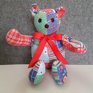 May include: A patchwork teddy bear with a red bow. The bear is made of different fabrics with floral and plaid patterns in shades of blue, red, green, and white.