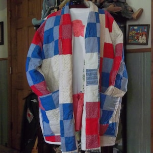 May include: A handmade patchwork jacket in red, white, and blue. The jacket features a quilted design with large square patches. It has two pockets and is hanging on a wooden door. The jacket is over a white shirt.