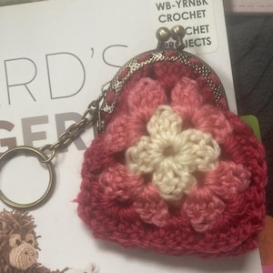 May include: A handmade crochet coin purse in shades of red and pink, featuring a cream-colored flower design in the center. The purse has a metal clasp and a keychain attachment. The background includes text from a book about crochet projects.