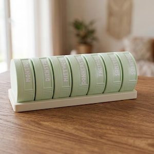 Può includere: Un portapillole settimanale verde chiaro con i giorni della settimana in bianco. L'organizzatore rettangolare ha sette scomparti individuali, ciascuno contrassegnato con un giorno della settimana.