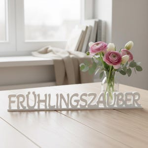 Könnte beinhalten: Auf einem Holztisch steht ein weißes Schild mit der Aufschrift "FRÜHLINGSZAUBER". Eine kleine Glasvase enthält rosa und weiße Blumen mit grünen Blättern. Der Hintergrund umfasst ein Fenster und Bücher.