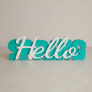 Könnte beinhalten: Ein dekoratives Schild mit dem Wort Hello in weißer Schreibschrift, das über dem Wort SPRING in fetten, türkisfarbenen Blockbuchstaben liegt. Das Objekt besteht aus zwei verschiedenen Materialschichten und erzeugt einen dreidimensionalen Effekt.