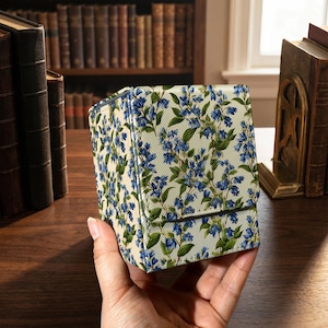 Puede incluir: Una caja cuadrada con estampado floral y cierre de solapa. La caja presenta un fondo crema con un patrón repetido de flores azules y hojas verdes. La caja está sujeta en la mano de una persona.