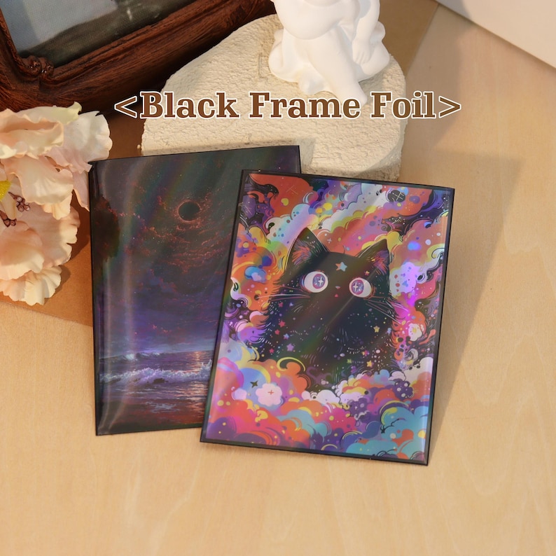 May include: Two rectangular prints with black frames labeled <Black Frame Foil>. One print features a dark ocean scene with a black sun, while the other displays a black cat surrounded by vibrant, multicolored clouds and stars with a foil finish.