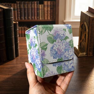 May include: A rectangular deck box featuring a vibrant floral pattern of blue, purple, and white hydrangea blossoms with green leaves against a white background. The box has a textured surface and a hinged lid that folds over the front panel.