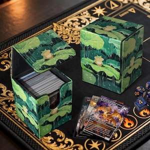 Puede incluir: Dos cajas de almacenamiento de cartas con un diseño de hojas verdes y ranas. Una caja está abierta, revelando una pila de cartas. Las cajas están sobre una superficie negra con detalles dorados, y se ven algunas cartas y dados.