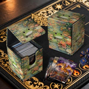 Puede incluir: Dos cajas de almacenamiento de cartas con un diseño de rana y nenúfares. Una caja está abierta, revelando una pila de cartas. Las cajas están sobre una superficie negra con detalles dorados. También se ven varias cartas y dados.