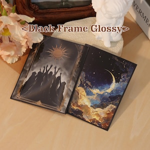 May include: A glossy black frame containing two rectangular art prints. The left print depicts silhouettes of figures reaching toward a sun in a dark sky. The right print shows a crescent moon surrounded by glowing clouds and stars in a night sky.
